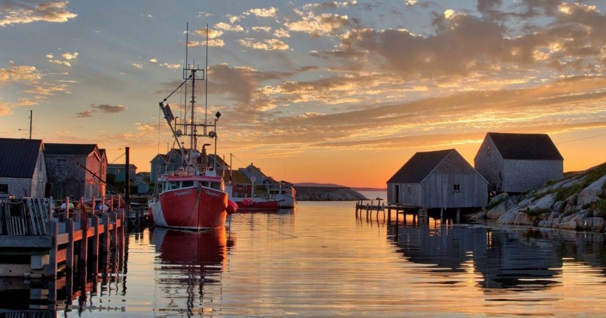 Peggy’s Cove, Nova Scotia Canada at sunset. A view out to the water with a fishing boat docked on the left and a wooden fishing shack on the right.