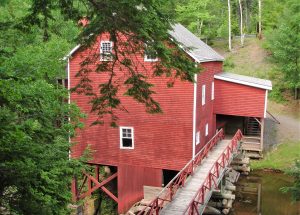 A red wooden building stands on stilts next to a bridged walkway by the water. Balmoral Grist Mill In,Tatamagouche Nova,Scotia Canada